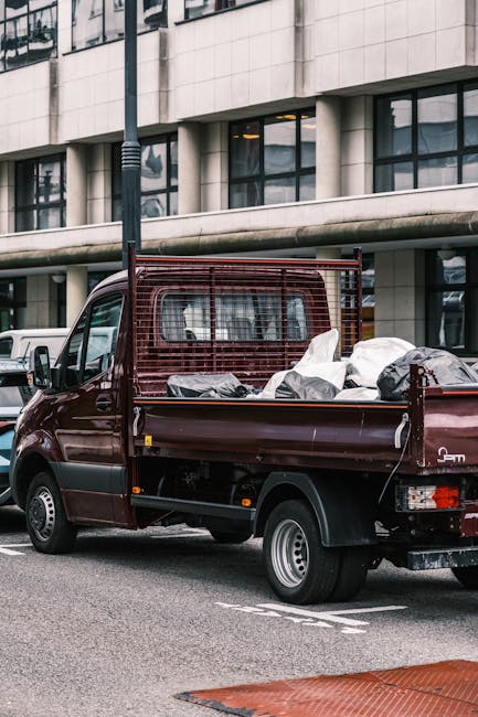 A small burgundy flatbed van parked on a city street, loaded with various household items and moving boxes covered with protective fabric and plastic wrap. The items include cardboard boxes, wrapped furniture pieces, and assorted packaging materials, positioned on the van's cargo area and secured with straps. The van is adjacent to a modern office building with large windows and concrete architecture, and a tall streetlamp is visible behind it. The scene depicts the preparation stage of home relocation or furniture transport, with the van belonging to Removal Companies Lambeth, supporting professional moving services in Lambeth SE1. The environment is well-lit, capturing the logistics involved in packing and loading household possessions for a house removal process.