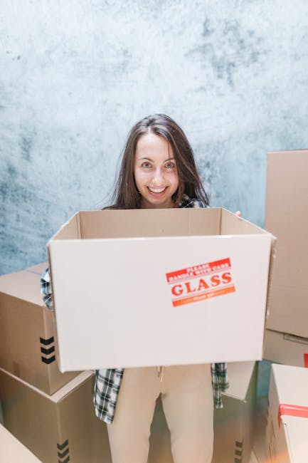A woman with long brown hair and a smile is holding a large empty cardboard box labeled 'FRAGILE GLASS' in red and white, standing indoors against a textured light blue wall. She is dressed in casual clothing, with a plaid shirt partially visible over a light top. Surrounding her are several other packed cardboard boxes of various sizes, some sealed with tape, positioned on the floor and stacked behind her. The boxes appear to be ready for a house move, reflecting packing and moving activities typical of home relocation services. The interior space is well-lit, and the scene captures the process of loading or packing items for a house removal by [COMPANY_NAME], illustrating a professional and organized approach to furniture transport and moving logistics.
