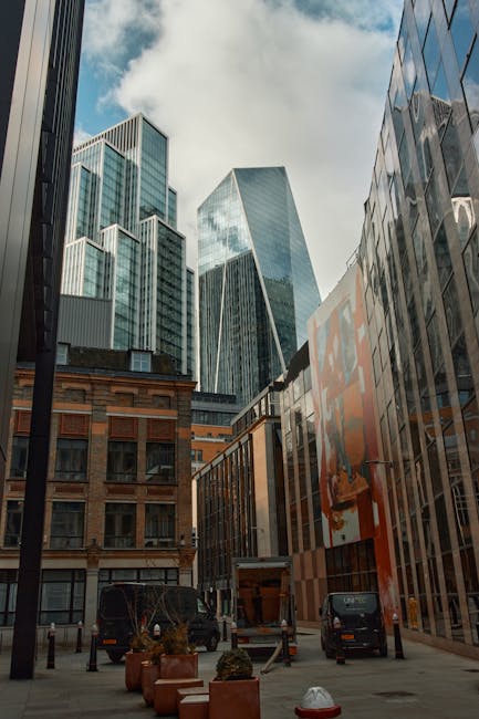 A small burgundy flatbed van parked on a city street, loaded with various household items and moving boxes covered with protective fabric and plastic wrap. The items include cardboard boxes, wrapped furniture pieces, and assorted packaging materials, positioned on the van's cargo area and secured with straps. The van is adjacent to a modern office building with large windows and concrete architecture, and a tall streetlamp is visible behind it. The scene depicts the preparation stage of home relocation or furniture transport, with the van belonging to Removal Companies Lambeth, supporting professional moving services in Lambeth SE1. The environment is well-lit, capturing the logistics involved in packing and loading household possessions for a house removal process.