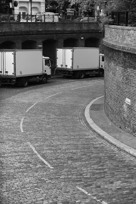 A photograph depicts a small, curved cobblestone street in an urban setting, with three white box trucks parked along the side, likely involved in a house removal or relocation service. The trucks are positioned parallel to a brick retaining wall on the right, which partly encloses the street and has a small white sign attached to it. In the background, there is a raised terrace or balcony with a metal railing and potted plants, possibly part of a residential building, indicating the area is a mixed-use or residential neighborhood. The street appears to be part of the loading area for furniture transport, with the trucks ready for loading or unloading household items, including cardboard boxes, wrapped furniture, or appliances, although these specific objects are not visible in the image. The environment is well-lit, suggesting daytime, and the scene captures the logistics process involved in home relocation handled by companies such as Removal Companies Lambeth, focusing on the loading process and transportation in a city setting.