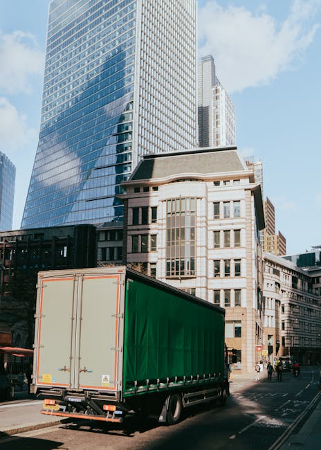 A large moving truck with a green cargo cover is parked on a city street in front of modern high-rise buildings with glass facades and older stone-fronted structures. The truck is positioned near the sidewalk, with its rear doors closed, likely prepared for loading or unloading furniture, packing boxes, and household appliances as part of a house removal process. The surrounding environment includes pedestrians walking nearby and other vehicles on the road. The clear sky and daylight highlight the urban environment, reflecting the city's busy and organized atmosphere. This scene illustrates the logistics involved in home relocation, supported by a professional moving service such as Removal Companies Lambeth, subtly emphasizing the careful handling of household items during furniture transport and packing and moving activities.