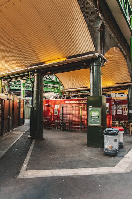 An indoor scene at Brixton Market featuring a section of the covered walkway with a curved, ribbed yellow ceiling supported by black metal columns. In the background, there is a red metal-framed market stall with signage and small windows, surrounded by other market structures and green scaffolding. On the right side of the image, a black trash bin and a red-lidded recycling bin are positioned near a black post with a green and white leaf logo. The floor is paved with dark asphalt, with visible parking or loading lines. The scene is well-lit with natural light filtering through the market area. This environment exemplifies an active market space suitable for home relocations or furniture transport involving loading and unloading activities, with visible market infrastructure conforming to moving and packing logistics managed by companies such as Removal Companies Lambeth.