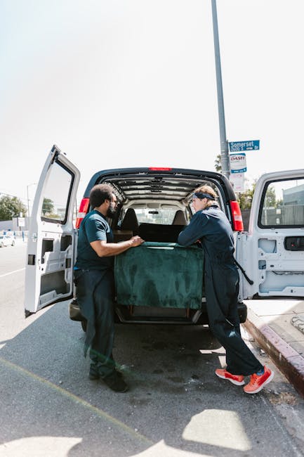 Two removal specialists from Removal Companies Lambeth are engaged in the loading process of a home relocation, lifting a large, green upholstered piece of furniture into the open back of a black cargo van. The interior of the van shows a spacious area suitable for furniture transport, with some visible packaging materials. Both workers are dressed in navy uniforms, wearing gloves, and are working together to carefully carry the item, ensuring proper handling during the packing and moving operation. The van is parked on a paved street near a curb, with a street sign and supporting pole visible, indicating the urban surroundings of Brixton Market in Lambeth. The scene is illuminated by natural daylight, highlighting the professional approach to a furniture transport service, emphasizing the logistical aspect of house removals and efficient loading procedures typically involved in relocation services.