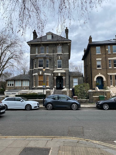 A three-storey Victorian-style residential building with a dark grey brick exterior, located on a street with other similar houses. The building has a central front door accessible via a short flight of steps, with tall bay windows on the ground and first floors, and a small dormer window in the roof. Two chimneys are visible on the roof. In front of the house, three parked cars are aligned along the street sidewalk— a white, a grey, and a black vehicle. Leafless trees with thin branches are seen on either side of the building, and the sky above is overcast with grey clouds. The street scene captures the typical urban setting for house removals and property transfers, with the house awaiting a move or ongoing packing and loading activities facilitated by Removal Companies Lambeth, as part of a house relocation or furniture transport process.