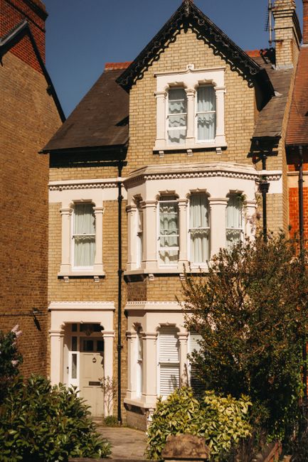 A three-storey Victorian-style residential building with a beige brick facade, white decorative window surrounds, and a dark, pitched roof. The ground floor features a white front door and windows with curtains, partially obscured by greenery and shrubbery in the front garden. The building is situated on a narrow street with neighboring brick houses visible on either side. This image is associated with house removals and relocation services, illustrating the type of property typical of a full house move by Removal Companies Lambeth, involving packing, loading, and transporting household items from this Brixton address to a new location in SE1.