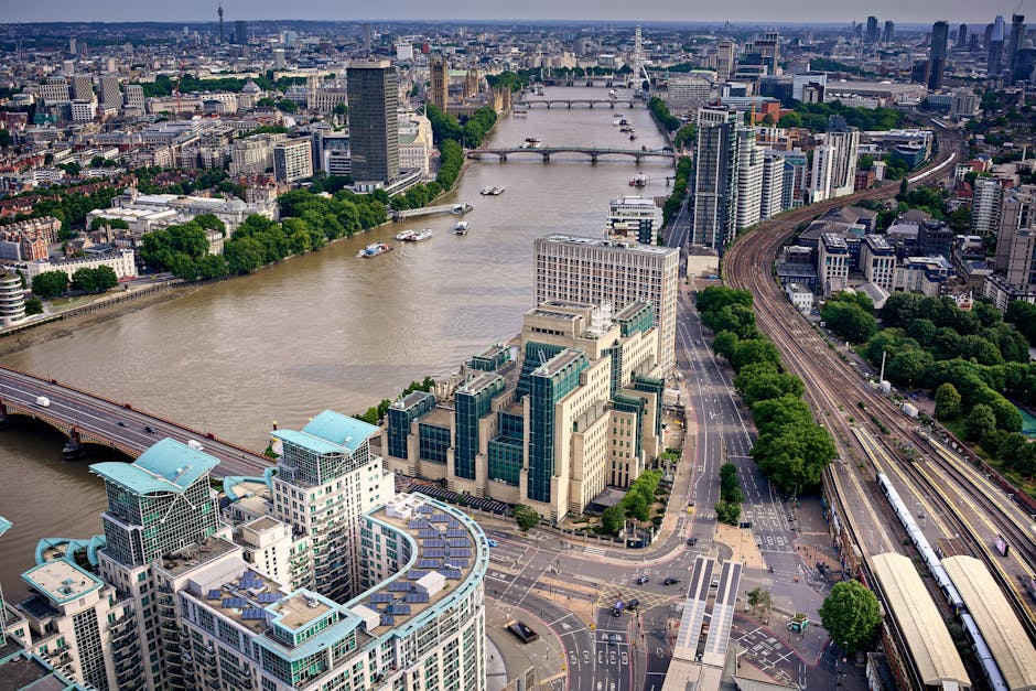 An aerial view of central London showing the River Thames with several boats and bridges crossing over it, surrounded by tall modern office buildings with glass exteriors and green rooftops. To the right, a railway line runs parallel to a tree-lined street, with a train visible on the tracks. On the left side of the image, a bridge connects buildings across the river, with a mix of commercial and residential structures along the riverbank. The scene captures a busy urban environment during daylight hours with overcast skies, illustrating the cityscape where house and office removals could involve moving furniture and belongings through the streets or via transport in a busy metropolitan area. The image is relevant to services such as removals and office relocations provided by Removal Companies Lambeth, highlighting the logistical environment for furniture transport and relocation planning in central London.