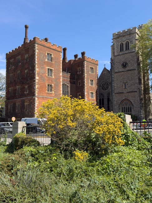 The image depicts a historic red brick building with multiple windows, situated behind a small, well-maintained garden area with yellow-flowering bushes and green foliage in the foreground. To the right, there is a stone church with a clock tower, arched windows, and crenellated roofline, indicating its Gothic architectural style. In front of the building and garden, a street with parked cars and a metal fence is visible, with some vehicles partially obscured by the greenery. The scene is captured on a clear day with blue skies, and ample natural light illuminates the structures and landscape. This setting aligns with a London neighbourhood often associated with professional house removals and local relocation services, such as those provided by Removal Companies Lambeth. The image highlights the combination of historic architecture and urban environment, relevant to home or office transport and packing processes involved in moving and relocation activities.