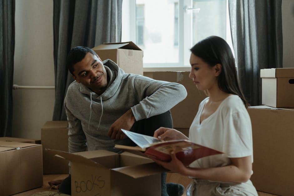 A young man and woman inside a room filled with cardboard moving boxes of various sizes, some labeled with handwritten notes such as 'BOOKS.' The man, wearing a grey hoodie, is seated on the floor and looking at the woman with a slight smile, while the woman, dressed in a white top, is holding an open book or folder and appears to be reading or discussing it. Behind them, there is a window with sheer curtains allowing natural light to illuminate the scene, and additional boxes are stacked on a windowsill and nearby surfaces. Visibility of packing materials such as plastic wrap or packing paper is not apparent, but the scene suggests an ongoing home relocation process managed by Removal Companies Lambeth. The setting indicates an interior environment prepared for moving or unpacking, with people engaged in organizing or boxing belongings as part of furniture transport or moving logistics activities.