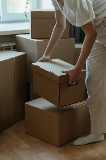 A person wearing light-colored clothing and white socks is shown indoors, carefully handling a medium-sized cardboard box during a home relocation process. The individual is placing the box onto a stack of similar cardboard boxes, which vary in size and are made from plain brown packaging material. Some boxes have labels, such as one marked 'Clothes,' indicating their contents. The scene takes place in a well-lit room with natural light coming through a window in the background. Additional cardboard boxes are visible nearby, suggesting a packing phase of a house move. The person’s hands grip the box firmly as they prepare to load or transport it, possibly involving furniture transport and moving logistics. Such activity is typical during packing and loading processes carried out by professionals like Removal Companies Lambeth, who assist with house removals and furniture transport within Lambeth flats and homes.