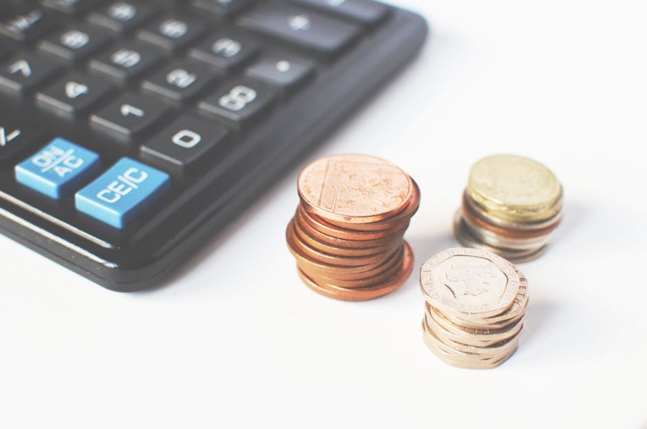 A close-up image displays a black calculator with blue and white keys, positioned next to three stacks of Euro coins in various denominations, including one-cent, five-cent, and ten-cent pieces, arranged on a white surface. The calculator's buttons are clearly visible, showcasing numerical and operational symbols, while the coins are neatly stacked, with some slightly tilted. The setting appears to be an indoor workspace, with the coins and calculator used for financial calculations related to house removal costs or moving logistics, as referenced on the page about cost comparison of house removals in SE24 Tulse Hill versus DIY options. The image represents financial aspects of home relocation services offered by Removal Companies Lambeth, emphasizing cost considerations and budget planning for house moves.