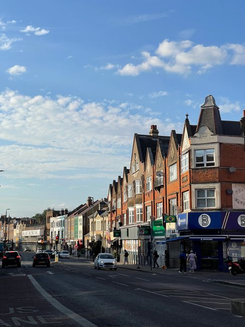 A street scene in an urban residential area showing a row of multi-story brick houses with pitched roofs and large windows. Several pedestrians are walking on the sidewalk, and streetcars or buses are visible at the bus stop. Vehicles, including cars and a van, are driving along the road, which is lined with shops and storefronts at ground level. The sky above is partly cloudy with patches of blue, indicating daytime. This image, associated with house removals and home relocation services, illustrates the typical environment where furniture transport and packing during a move are conducted, reflecting the context of comparing professional removals versus DIY options for residential moves in the SE24 Tulse Hill area, as detailed by Removal Companies Lambeth.
