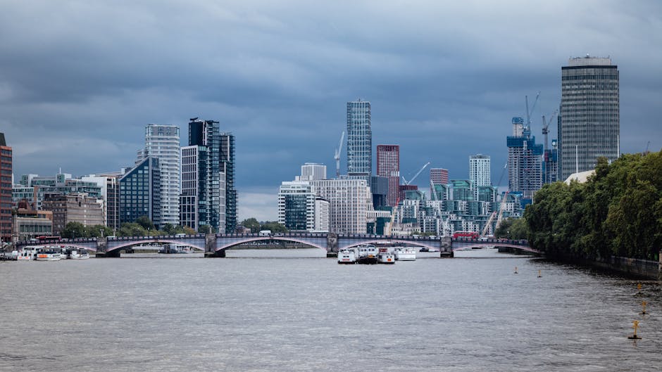 A wide view of the River Thames in central London, with several white boats and larger vessels moored along the water. In the background, there is a dense cluster of modern high-rise buildings featuring glass facades, some under construction with cranes visible. To the right, there is a green, tree-lined area along the riverbank. The sky is overcast with dark clouds, casting a subdued light over the scene. This image captures an urban landscape typical of a home relocation or furniture transport process in the Kennington SE11 area, with buildings indicating a busy city environment suitable for house removals by Removal Companies Lambeth, emphasizing the logistics of loading and moving furniture through the cityscape.