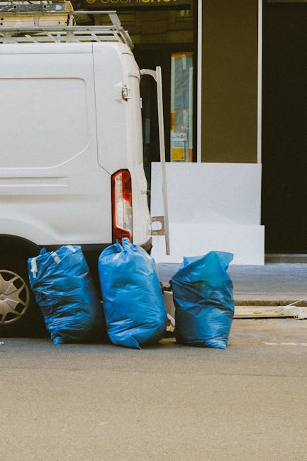 A white commercial van parked on a city street with its rear doors closed, partially visible in the image. Four large blue plastic rubbish bags, filled and knotted at the top, are placed on the pavement next to the van's rear wheel, indicating waste disposal or preparation for removal. Nearby on the sidewalk, there is a white support panel leaning against the building's entrance, which features a glass door and window, suggesting a residential or commercial property. The scene is illuminated by natural daylight, with the setting likely involved in home or office relocation activities, as indicated by the context from Removal Companies Lambeth. This image reflects the process of managing waste materials associated with furniture, packaging, or items scheduled for removal during a house move or clearance, aligning with services related to house removals and logistical support in Lambeth.