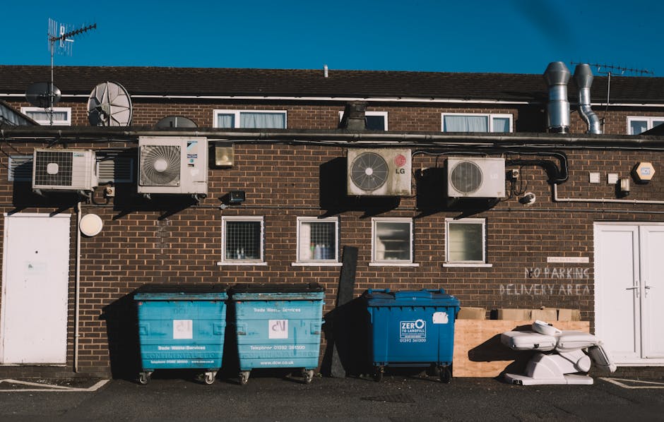 A white commercial van parked on a city street with its rear doors closed, partially visible in the image. Four large blue plastic rubbish bags, filled and knotted at the top, are placed on the pavement next to the van's rear wheel, indicating waste disposal or preparation for removal. Nearby on the sidewalk, there is a white support panel leaning against the building's entrance, which features a glass door and window, suggesting a residential or commercial property. The scene is illuminated by natural daylight, with the setting likely involved in home or office relocation activities, as indicated by the context from Removal Companies Lambeth. This image reflects the process of managing waste materials associated with furniture, packaging, or items scheduled for removal during a house move or clearance, aligning with services related to house removals and logistical support in Lambeth.