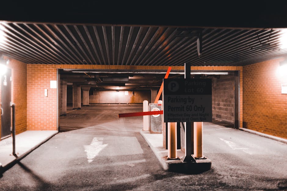 The image depicts an underground parking garage entrance with a brick wall on the left and right sides, and a black corrugated ceiling overhead. In the foreground, there is a designated parking control post with a sign displaying parking regulations, including a 'Parking Lot 60' permit requirement, positioned next to a barrier arm gate that is lifted, allowing vehicle access. The pavement in front of the gate is marked with white directional arrows, guiding vehicles into the garage. Inside, the parking area is dimly lit with concrete support columns and an unevenly illuminated space that extends into the background. Near the entrance, a small platform supports the control post and barrier arm, indicating an organized vehicle entry point. This scene reflects the logistical setup associated with house removals and moving services, emphasizing the importance of parking permissions, access control, and efficient vehicle loading for successful relocation projects, as highlighted on the 'Lambeth Council parking permits for house removals' page. Removal Companies Lambeth may utilize such parking facilities for home relocation and furniture transport operations.