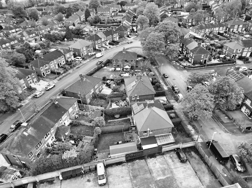 An aerial black-and-white photograph showing a residential area with closely spaced houses, many with gardens and pitched roofs. Several cars are parked along the narrow streets that wind through the neighbourhood. In the foreground, a house is being prepared for a home relocation, with visible packing materials such as cardboard boxes, plastic wrapping, and furniture covers placed around the house and on the driveway. Removal Companies Lambeth is managing the process, with furniture being carried by movers and transported onto a loading area near a van. The scene captures the methodical loading process typical of house removals, with tools like trolleys and straps possibly in use, although not clearly visible. The environment is well-lit, and the overall setting demonstrates organized logistics involved in furniture transport and packing during a house move, supporting the themes of house removals and relocation services related to Lambeth Council parking permits for house removals.