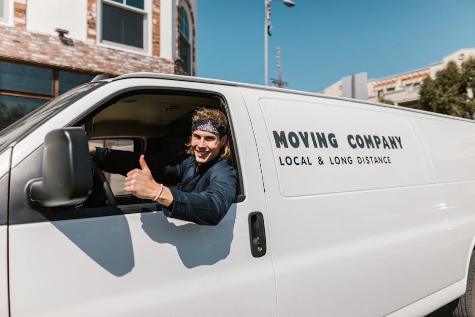 A young male mover with long, wavy hair, wearing a black headband and a dark blue shirt, is sitting in the driver's seat of a white removal van with the company name 'Moving Company' and the descripti
