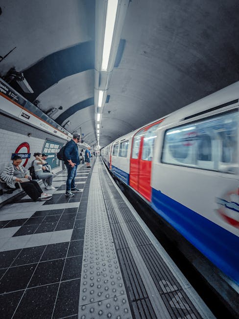 Inside a London underground station, the platform features a group of passengers sitting and waiting for a train, with some standing near the edge. The platform has black and grey checkered flooring w