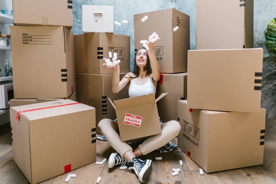 A young woman with curly black hair, wearing a white t-shirt and jeans, is seated on a wooden floor surrounded by numerous cardboard moving boxes of various sizes. She is smiling and holding a large c